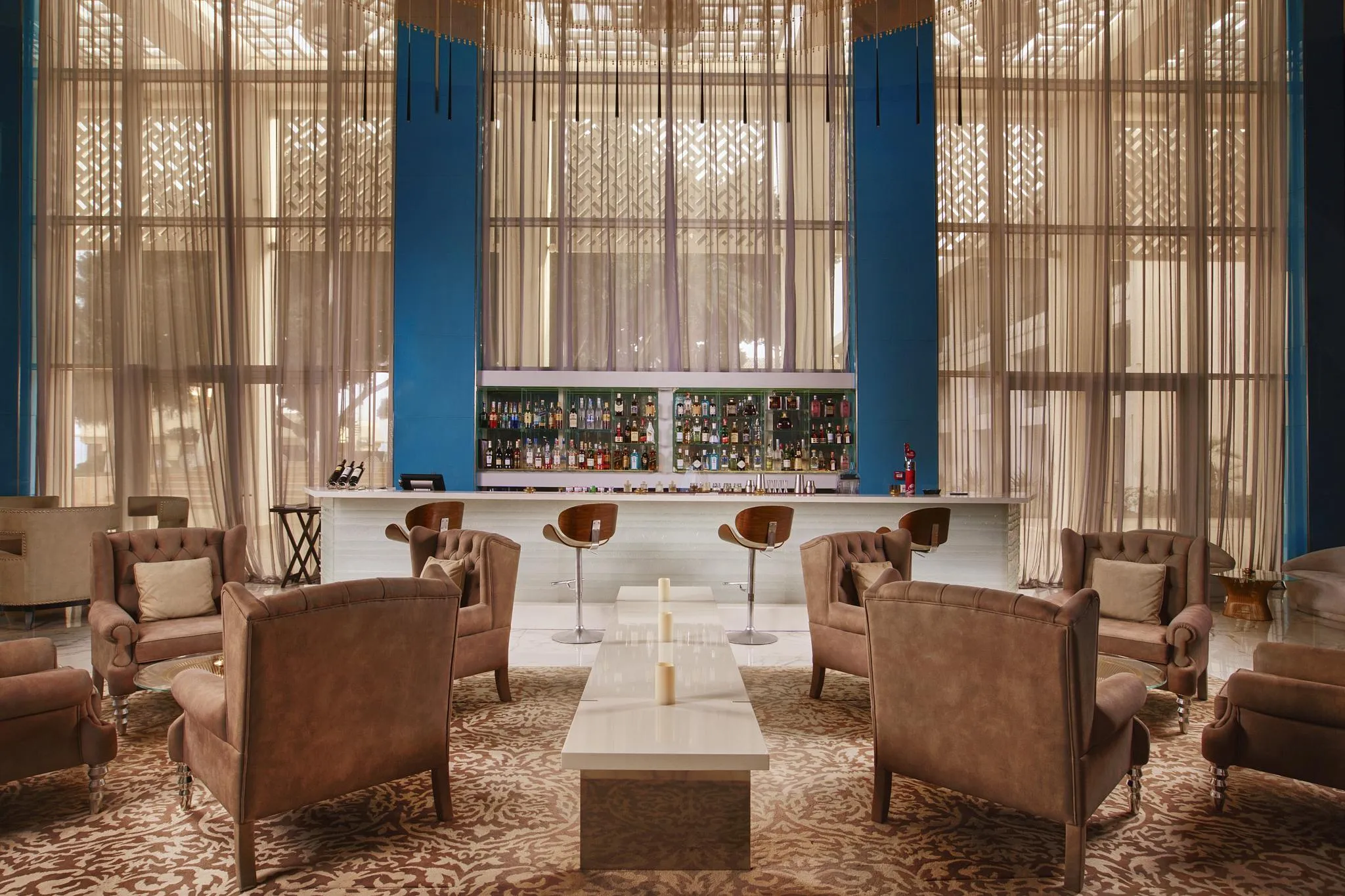 Un salon avec des fauteuils en cuir autour d'une table basse, face à un bar avec des étagères remplies de bouteilles, derrière de grandes fenêtres aux rideaux transparents