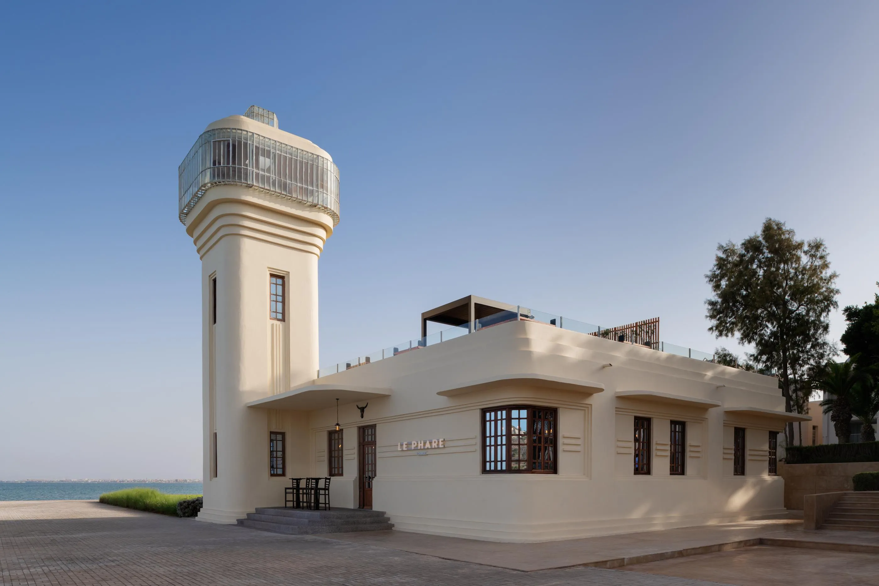 Un phare blanc avec une tour carrée et une plateforme d'observation vitrée, situé près de la mer, avec un bâtiment bas adjacent portant l'enseigne "LE PHARE"