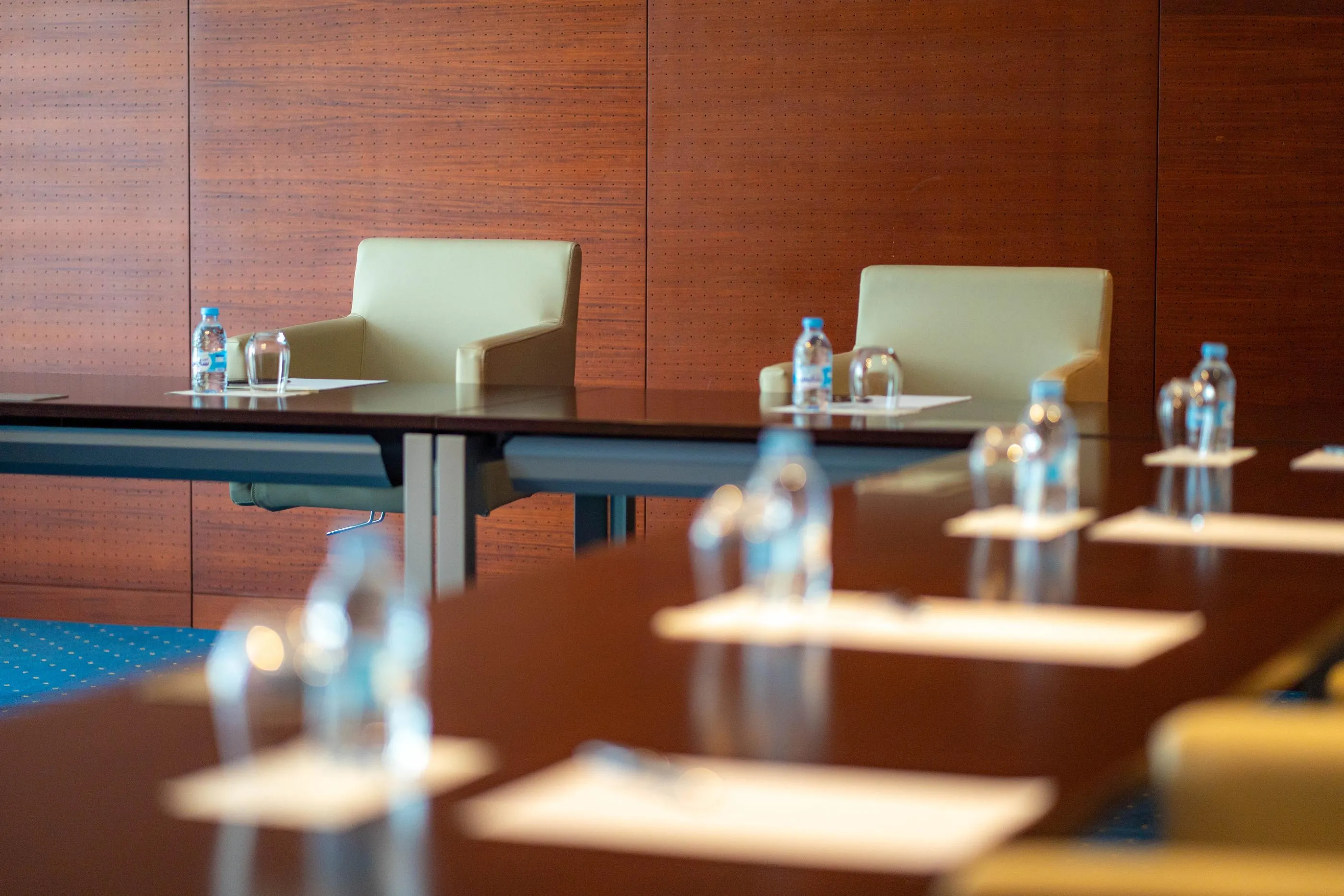 Une rangée de tables de conférence avec des bouteilles d'eau et des verres devant des chaises vides dans une salle aux murs en bois