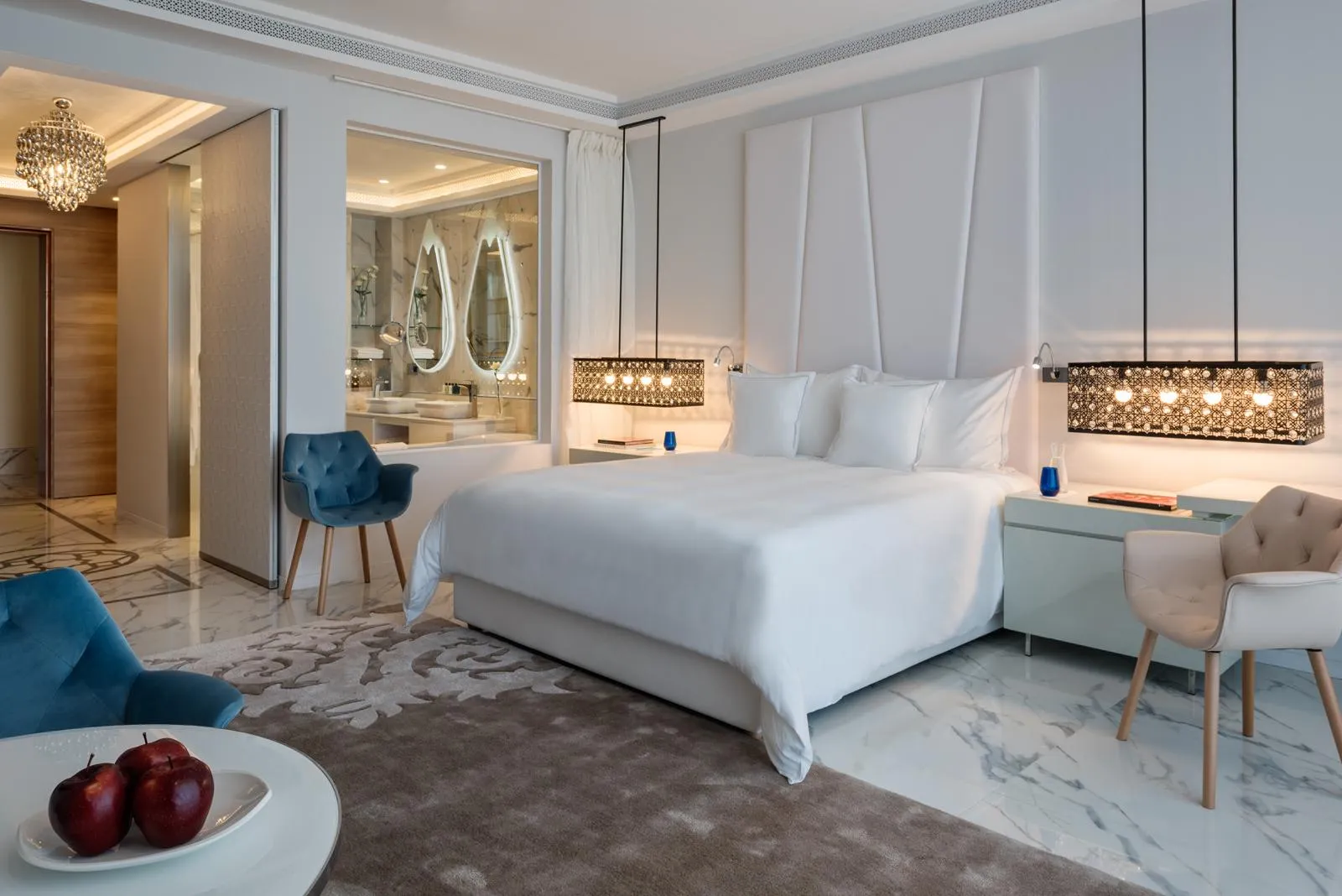 Chambre luxueuse avec lit blanc, deux fauteuils, table de chevet et vue sur une salle de bain avec miroirs ovales