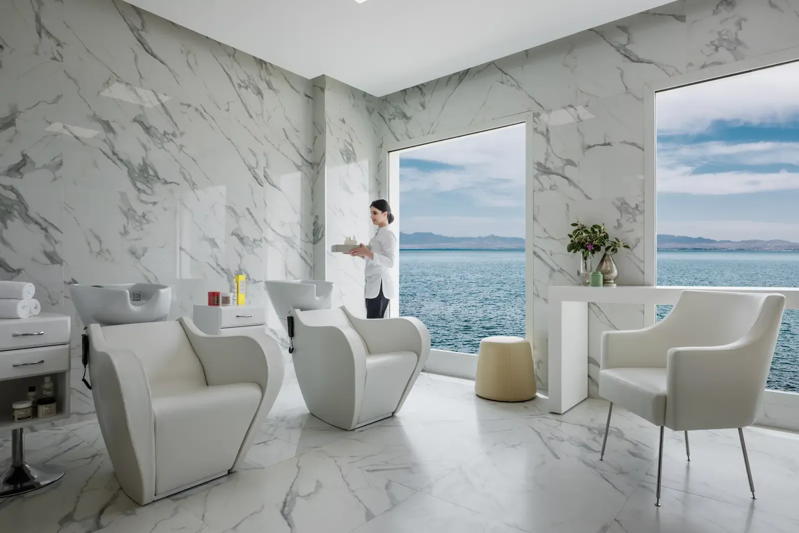 Salon de coiffure luxueux avec vue sur la mer, une personne tenant un plateau près des fauteuils de coiffure