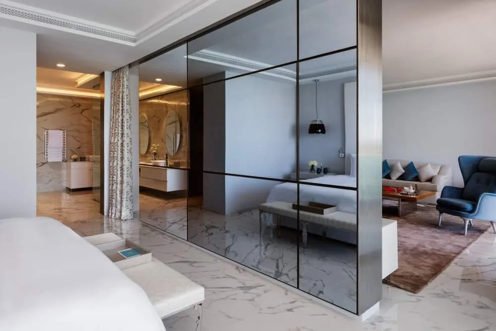 Intérieur moderne avec un miroir séparant une chambre à coucher et un salon, montrant des meubles blancs et un coin salon avec canapé et fauteuil