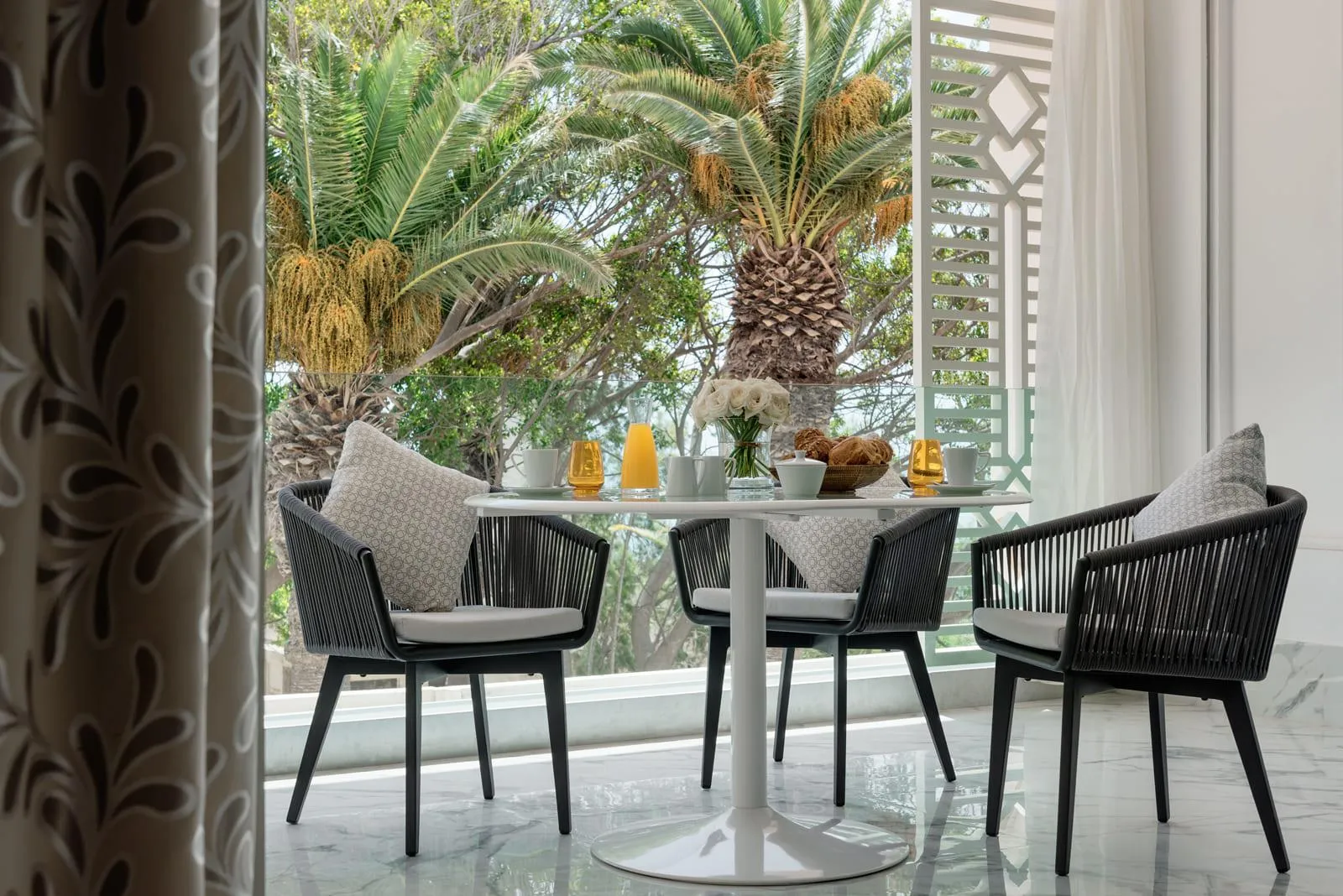 Table ronde avec quatre chaises en rotin noir et coussins gris, vue depuis une porte ouverte, avec des plantes et des éléments décoratifs sur la table en arrière-plan