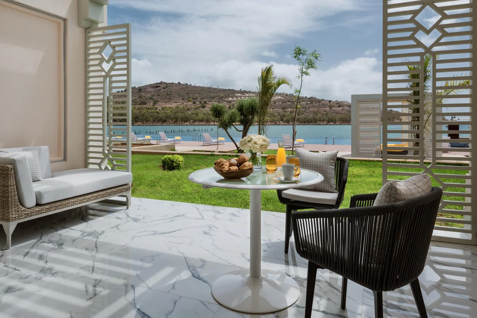 Terrasse avec table, chaises et canapé donnant sur une pelouse et un lac avec des transats et des montagnes en arrière-plan