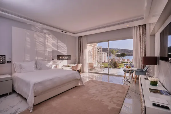 Chambre d'hôtel moderne avec un grand lit, une table et des chaises près d'une porte vitrée ouvrant sur une terrasse avec vue sur l'eau