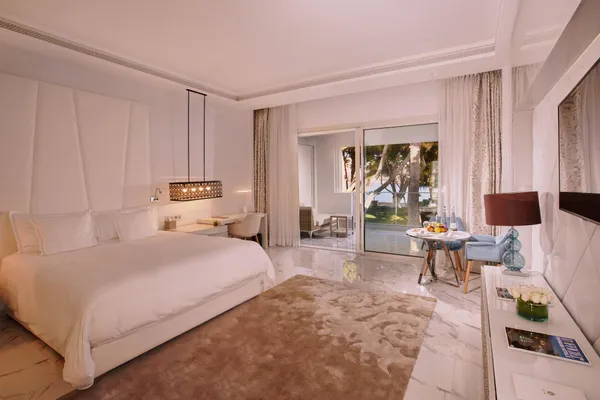 Chambre d'hôtel luxueuse avec un grand lit, une table avec des fruits et des bouteilles d'eau, et une porte-fenêtre donnant sur une terrasse avec vue sur des palmiers