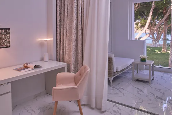 Bureau blanc avec chaise rose, lampe allumée, magazines et décor floral près d'une fenêtre donnant sur un jardin avec des arbres