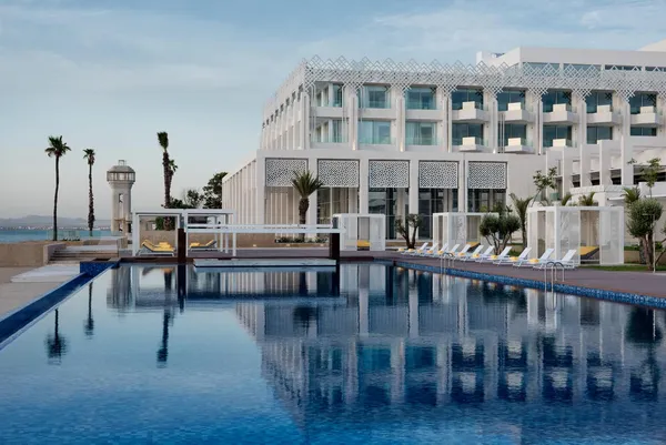 Piscine à débordement devant un hôtel moderne avec des transats et des palmiers, vue sur la mer en arrière-plan