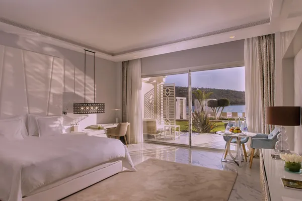 Chambre luxueuse avec lit blanc, table de travail, et vue sur une terrasse et un paysage extérieur depuis de grandes baies vitrées
