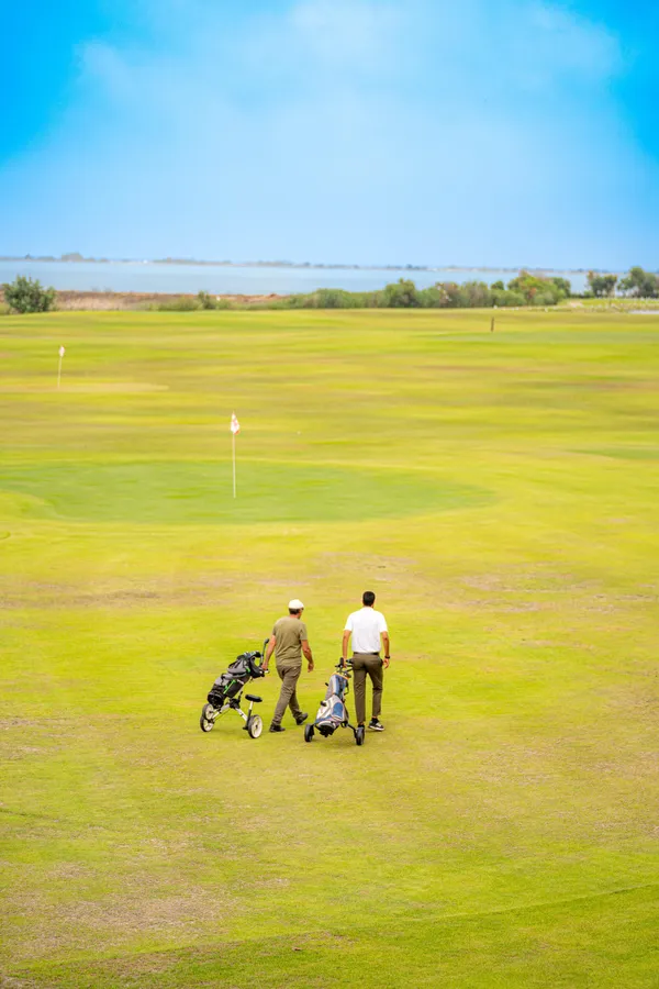 Deux personnes avec des chariots de golf marchent sur un green de golf avec un drapeau visible au loin et un plan d'eau à l'arrière-plan