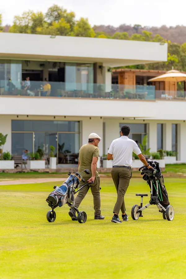 Deux hommes marchent sur un terrain de golf avec des sacs de clubs, devant un bâtiment moderne aux grandes baies vitrées