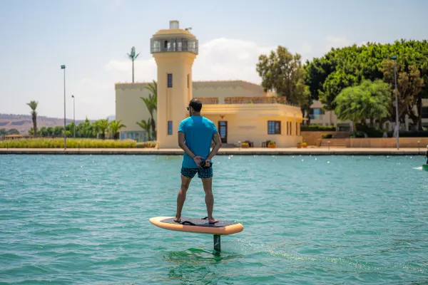 Un homme debout sur une planche de paddle dans une étendue d'eau, avec un bâtiment jaune en arrière-plan