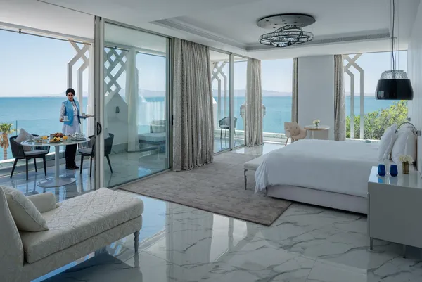 Chambre d'hôtel luxueuse avec vue sur la mer, un lit, un canapé, une table à manger et une personne servant un plateau